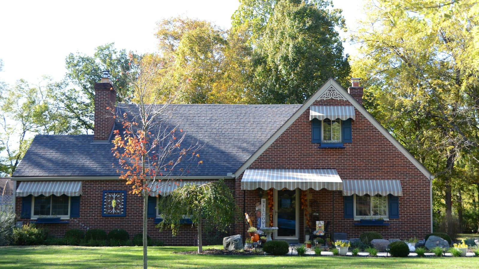 Red brick Cape Cod-style home with gray asphalt shingle roof, striped awnings, wide green lawn and autumn trees